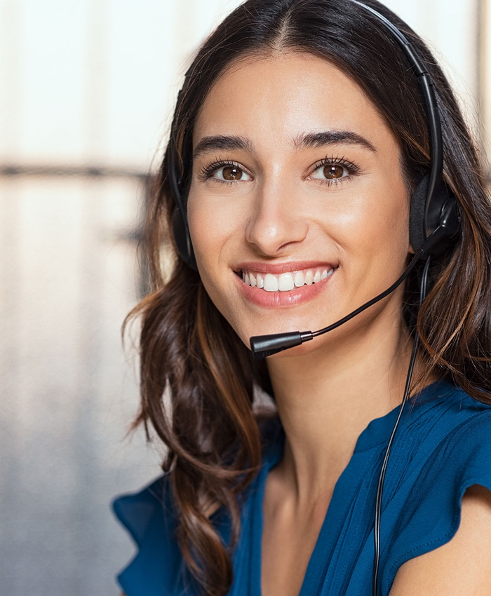 Eine Frau mit langen braunen Haaren, die ein Headset und ein blaues Oberteil trägt, lächelt in einer hellen Innenumgebung in die Kamera.
