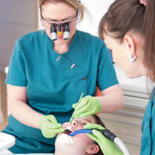 Vesna Radosevic und eine Helferin in blaugrünen Kitteln und grünen Handschuhen behandeln einer Patientin mit zahnärztlichen Werkzeugen und Absaugung, während einer eine Lupenbrille trägt. Der Patient liegt auf dem Rücken und hat den Mund geöffnet.