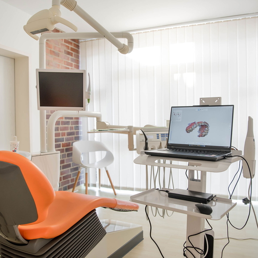 Bezahlgungszimmer mit einem orangefarbenen Behandlungsstuhl, einem an der Decke montierten Monitor und einem Laptop, der auf einem mobilen Wagen einen 3D-Zahnscan anzeigt. Der Raum ist hell, mit vertikalen Jalousien und einer Akzentwand aus Backstein.