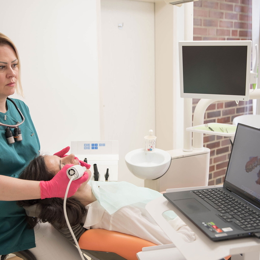 Zahnärztin Vesna Radosevic mit rosafarbenen Handschuhen benutzt ein Scangerät im Mund einer Patientin, während dieser sich in einem Zahnarztstuhl zurücklehnt. Ein Laptop und ein Monitor zeigen in der Nähe zahnmedizinische Bilder an.