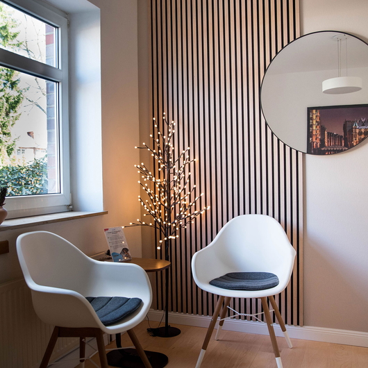 Gemütliches Wartezimmer mit zwei weißen Stühlen und grauen Kissen, einem kleinen runden Holztisch, einem dekorativen Baum mit Lichterketten, einem großen runden Spiegel und einem Fenster, das natürliches Licht hereinlässt.