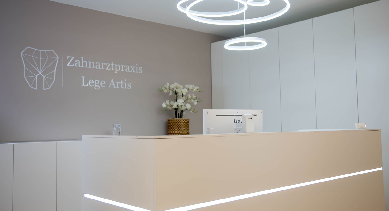 Moderner Empfangstresen mit einem schlichten weißen Schreibtisch, minimalistischem Dekor, einem Computermonitor und weißen Schränken. Die Wand zeigt das Logo der Zahnarztpraxis Lege Artis mit einem geometrisches Zahnlogo.