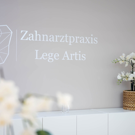 Minimalistisches Interieur der Zahnarztpraxis mit einem geometrischen Zahnlogo und dem Schriftzug Zahnarztpraxis Lege Artis an einer hellgrauen Wand, weißen Möbeln und einer weißen Orchideenpflanze auf einem Tresen.