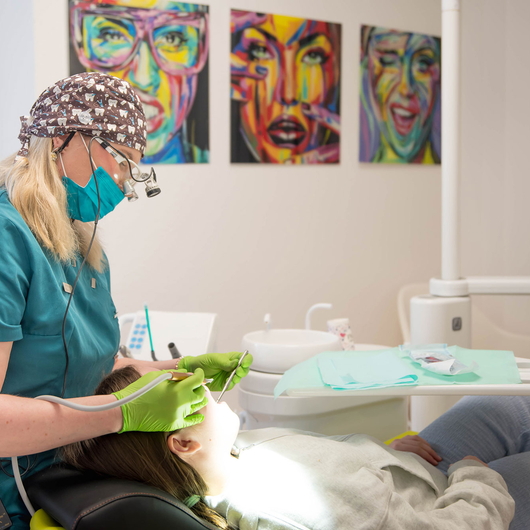 Zahnärztin Vesna Radosevic mit Maske, Handschuhen und Lupenbrille untersucht den Mund einer Patientin in einem hellen Behandlungsraum. An der Wand hinter ihnen sind bunte, abstrakte Porträts zu sehen.