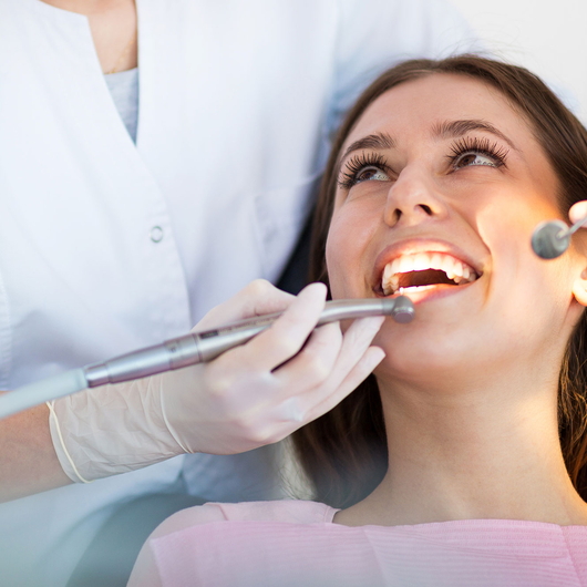 Eine Frau sitzt in einem Zahnarztstuhl und lächelt mit offenem Mund, während eine Zahnärztin mit weißen Handschuhen während einer zahnärztlichen Untersuchung Instrumente an ihre Zähne hält.