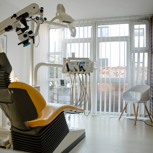 Moderne Behandlungsraum mit einem gelben Zahnarztstuhl, darüber hängenden zahnmedizinischen Geräten, hellem Tageslicht durch große Fenster mit Jalousien und einem weißen Stuhl in der Ecke.