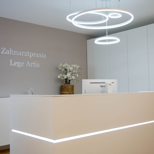 Moderner Empfangstresen mit einem schlichten weißen Schreibtisch, minimalistischem Dekor, einem Computermonitor und weißen Schränken. Die Wand zeigt das Logo der Zahnarztpraxis Lege Artis mit einem geometrisches Zahnlogo.