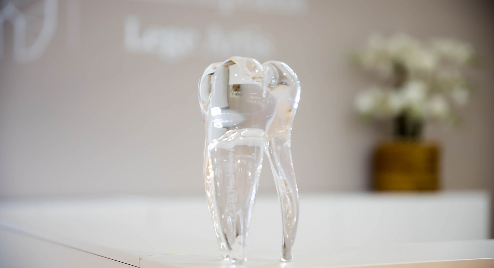 Eine klare Glasskulptur in Form eines Zahns steht auf einer weißen Fläche, vor einem unscharfen Hintergrund, der eine Topfpflanze und das unscharfe Logo an einer Wand zeigt.